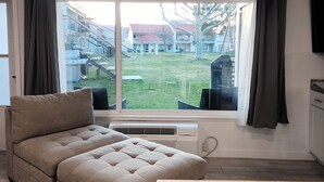 Ein Wohnzimmer mit einer Couch, einem Esstisch, einem großen Fenster mit Vorhängen und Blick auf ein Gebäude und einen Baum.