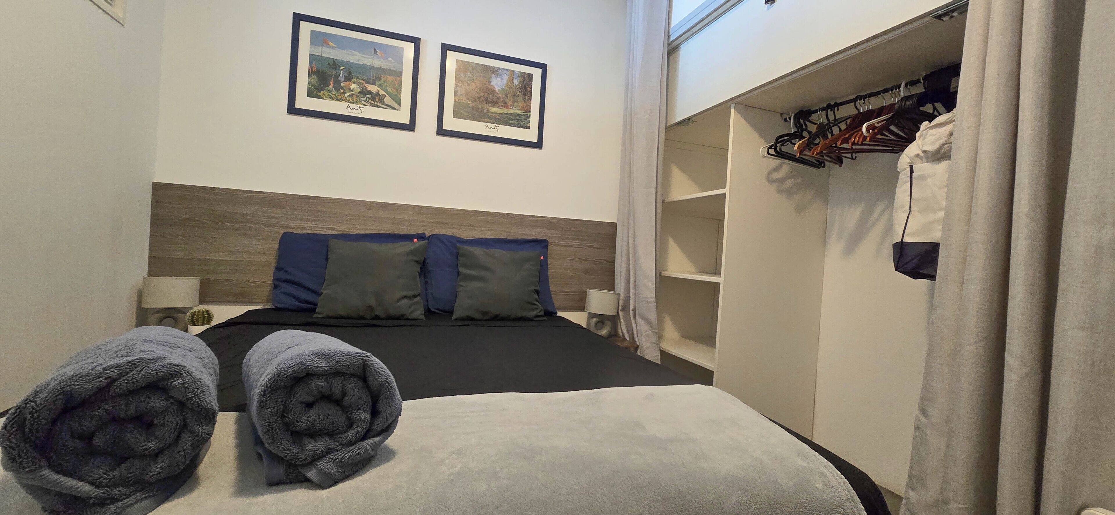 Ein Hotelzimmer mit einem Bett, zwei gerahmten Bildern an der Wand, einem Kleiderschrank mit Kleidung und zwei aufgerollten Handtüchern auf dem Bett.