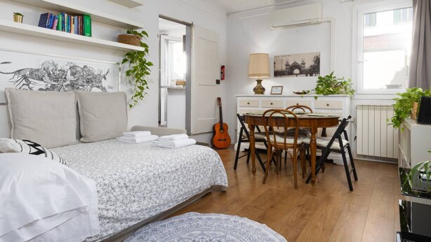 Quarto com cama, mesa de cabeceira e um violão. Há um espaço de jantar com mesa e cadeiras, e uma janela com cortinas.