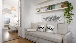 Uma sala de estar moderna com um sofá bege, uma parede branca com uma obra de arte emoldurada e uma estante branca com livros e objetos decorativos.