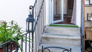 Uma escadaria com corrimão de metal leva a uma porta, cercada por plantas em vasos e um candeeiro.