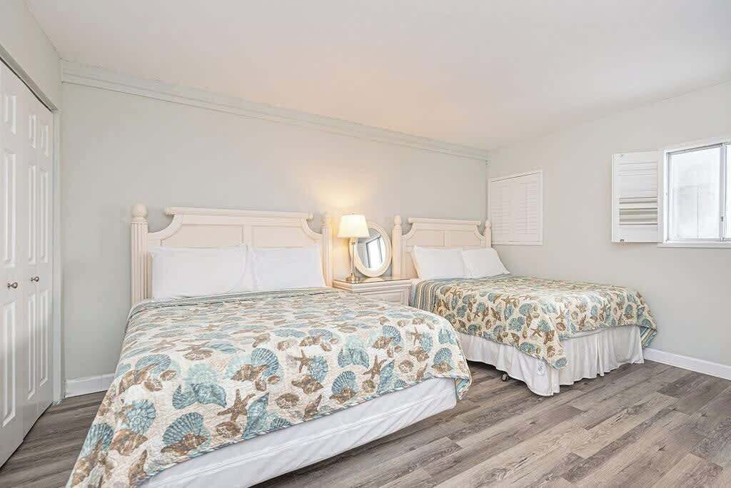 A bedroom with two beds, a nightstand with a lamp, a mirror, and a window with shutters.