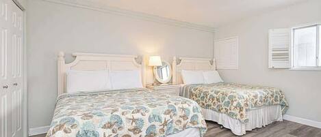 A bedroom with two beds, a nightstand with a lamp, a mirror, and a window with shutters.