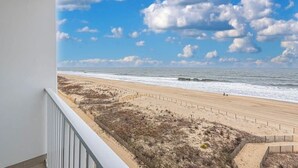 A beach view from a balcony with a railing, sandy beach, ocean, and a clear sky with clouds.