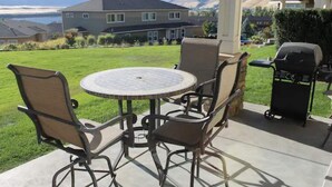 Eine Terrasse mit rundem Tisch, vier Stühlen, einem Grill und Blick auf ein Haus und Berge.