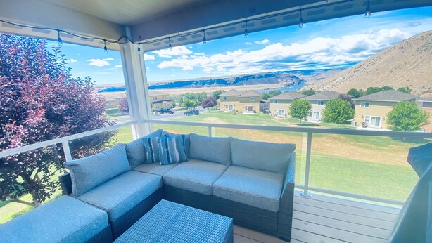 Ein überdachter Balkon mit einer Couch und Hocker, Blick auf eine Wohngegend und einen klaren Himmel.