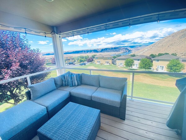 A covered balcony with a sofa and ottoman, a view of a residential area, and a clear sky.