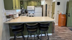 A kitchen with green cabinets, a white refrigerator, a microwave, and a sink. There are three black bar stools at the island. - Cozy Downtown Sitka Home, Car Rental Option (Sitka)