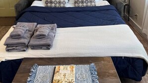A neatly made bed with a dark blue and white duvet, grey pillows, and folded towels on a white pillowcase. There is a wooden table with a woven placemat and a book titled "Compostable Notes." - Cozy Downtown Sitka Home, Car Rental Option (Sitka)