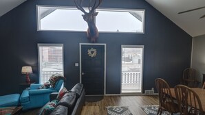 A living room with a high ceiling, a mounted deer head, a blue sofa, a dining table with chairs, and a door with a wreath. - Red River Retreat (Red River)