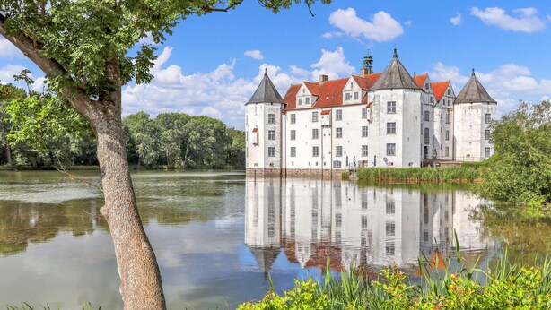 Un grand bâtiment historique doté de nombreuses tours et entouré de douves, le tout noyé dans une végétation luxuriante et bordé d’une étendue d’eau paisible.