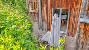 Un bâtiment rustique en bois, doté d’un espace extérieur couvert avec des sièges et d’une fenêtre donnant sur un jardin.