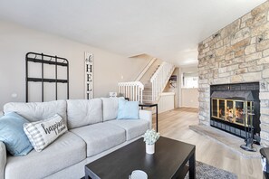 Una sala de estar moderna con chimenea de piedra, un sofá gris con almohadones decorativos, una mesa de centro negra y una escalera que sube a otro nivel.