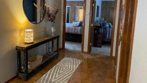 A hallway with a console table, a mirror, and a rug leading to a bedroom with a bed and a nightstand. - Doney Creek Cabin (Lakehead-Lakeshore)