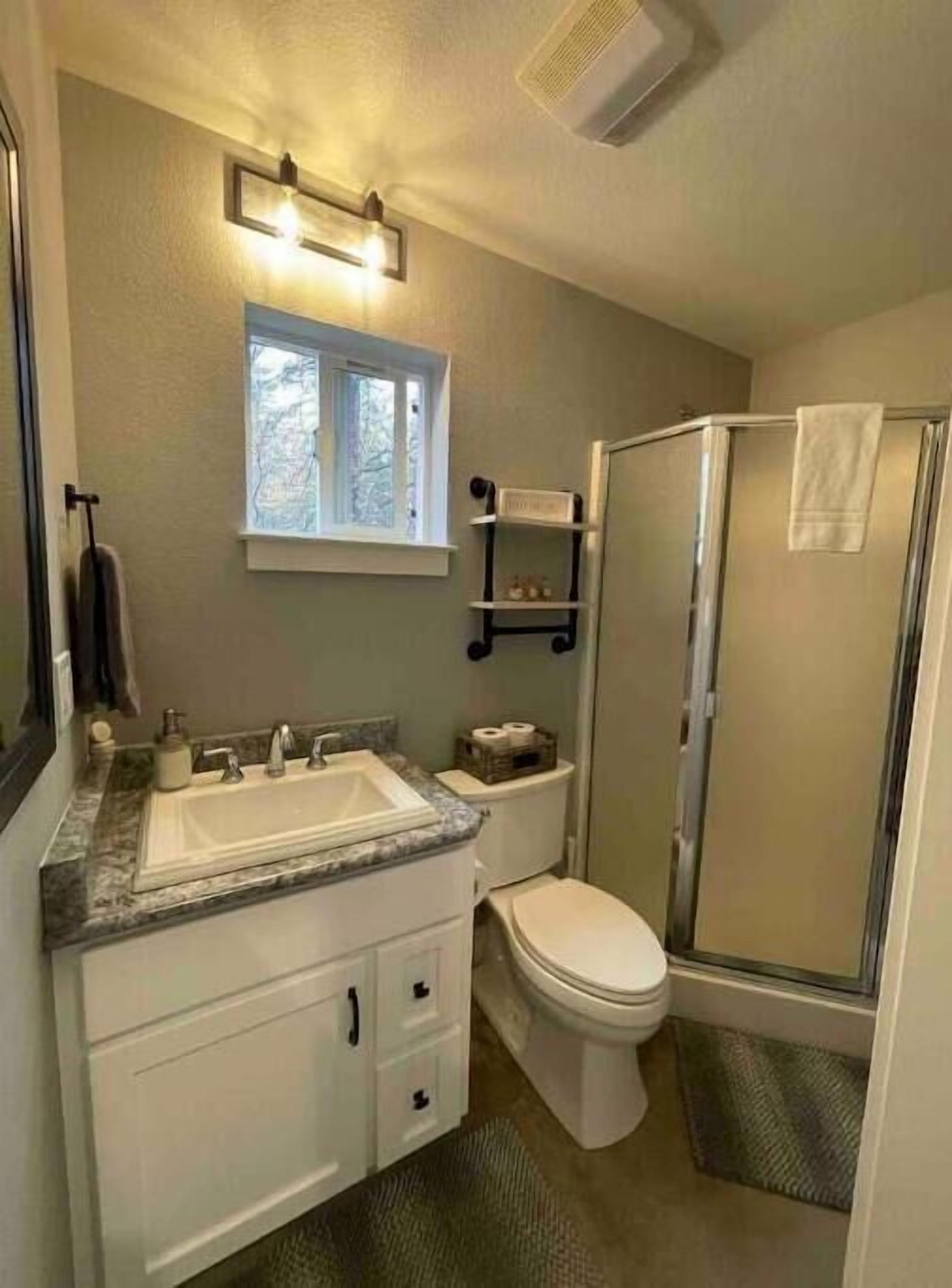 Une salle de bain avec un lavabo, des toilettes et une douche. Il y a une fenêtre au-dessus du lavabo et une serviette est accrochée à la porte de la douche.