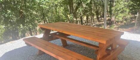 Una mesa de picnic de madera con bancos, ubicada sobre grava y rodeada de árboles.