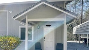 A house with a covered porch, two chairs, a door, and a small garden. - Doney Creek Studio (Lakehead-Lakeshore)