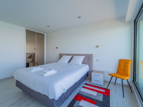 A modern hotel room with a large bed, a nightstand, a chair, and a view through a large window.