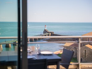 A coastal view from a window overlooking a pier leading to a lighthouse, with a table and chairs set for dining.