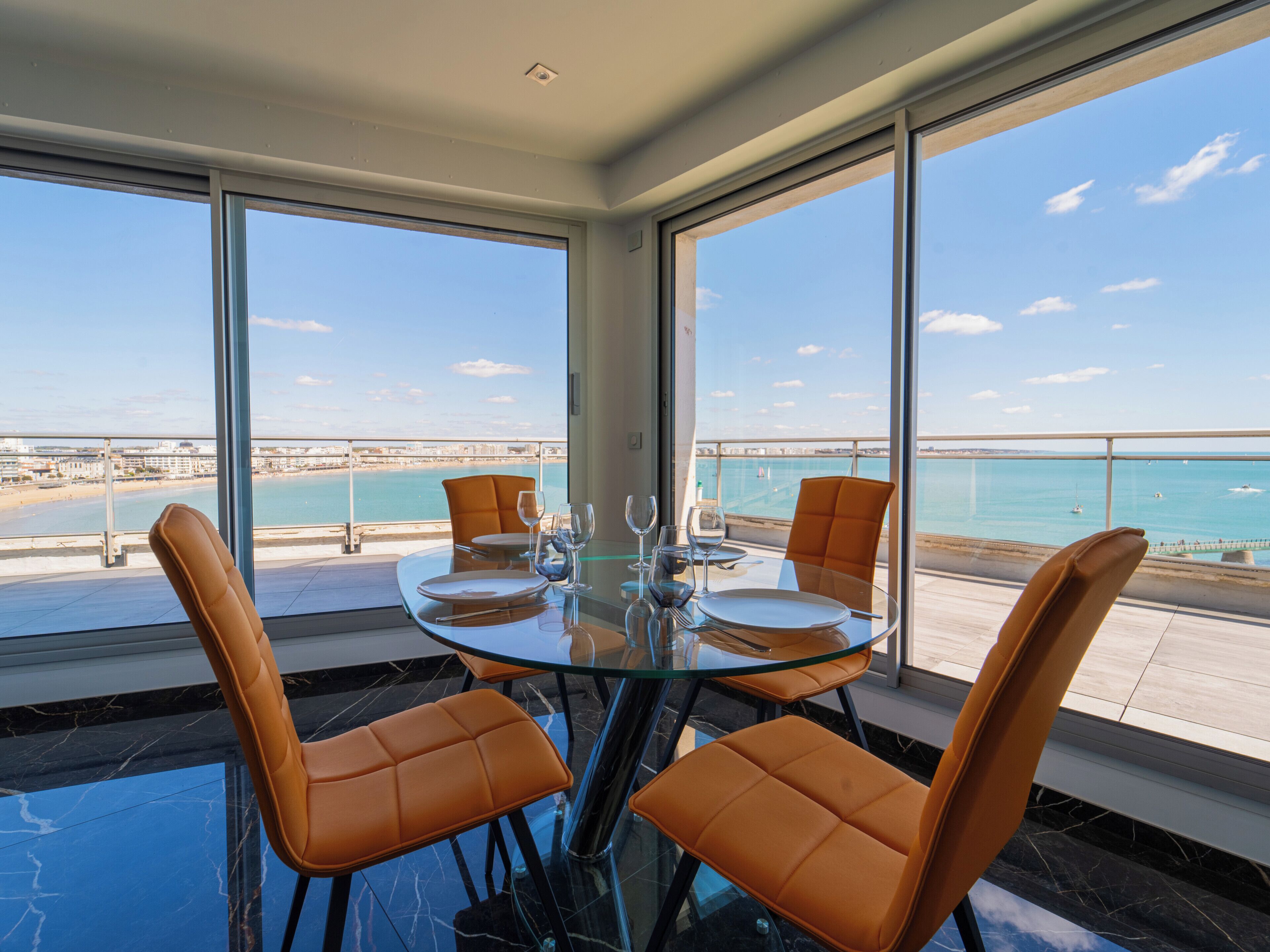 A dining area with a glass table and orange chairs, overlooking a beach and the sea.