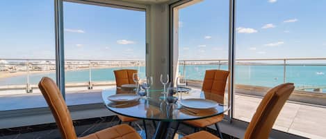 A dining area with a glass table and orange chairs, overlooking a beach and the sea.
