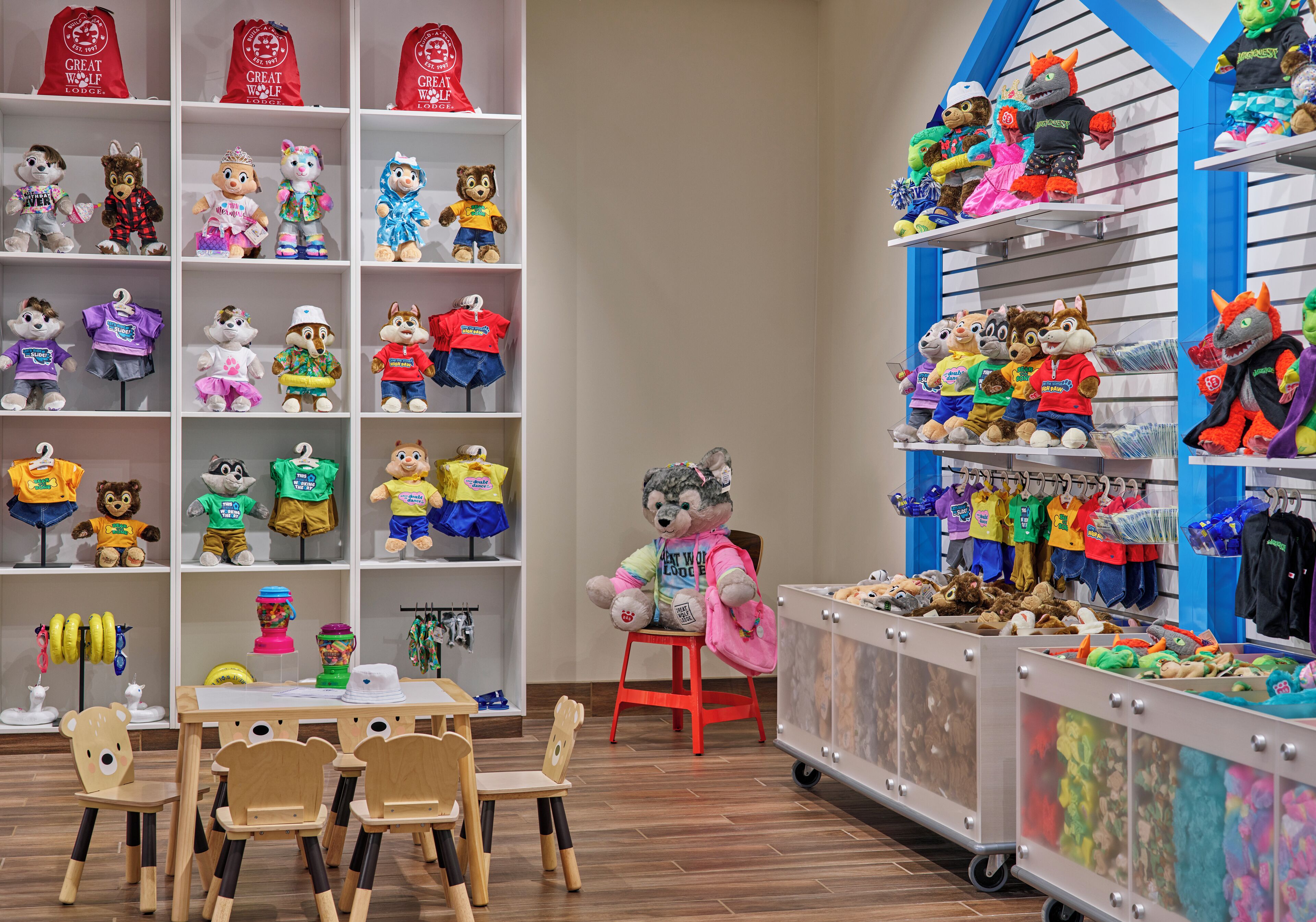 Een kinderspeelruimte met pluchen speelgoed op rekken, een tafel met stoelen en een groot knuffeldier in het midden.