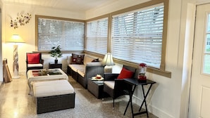 A cozy corner seating area with a sofa, ottoman, and a small table, featuring a window with blinds and a lamp.