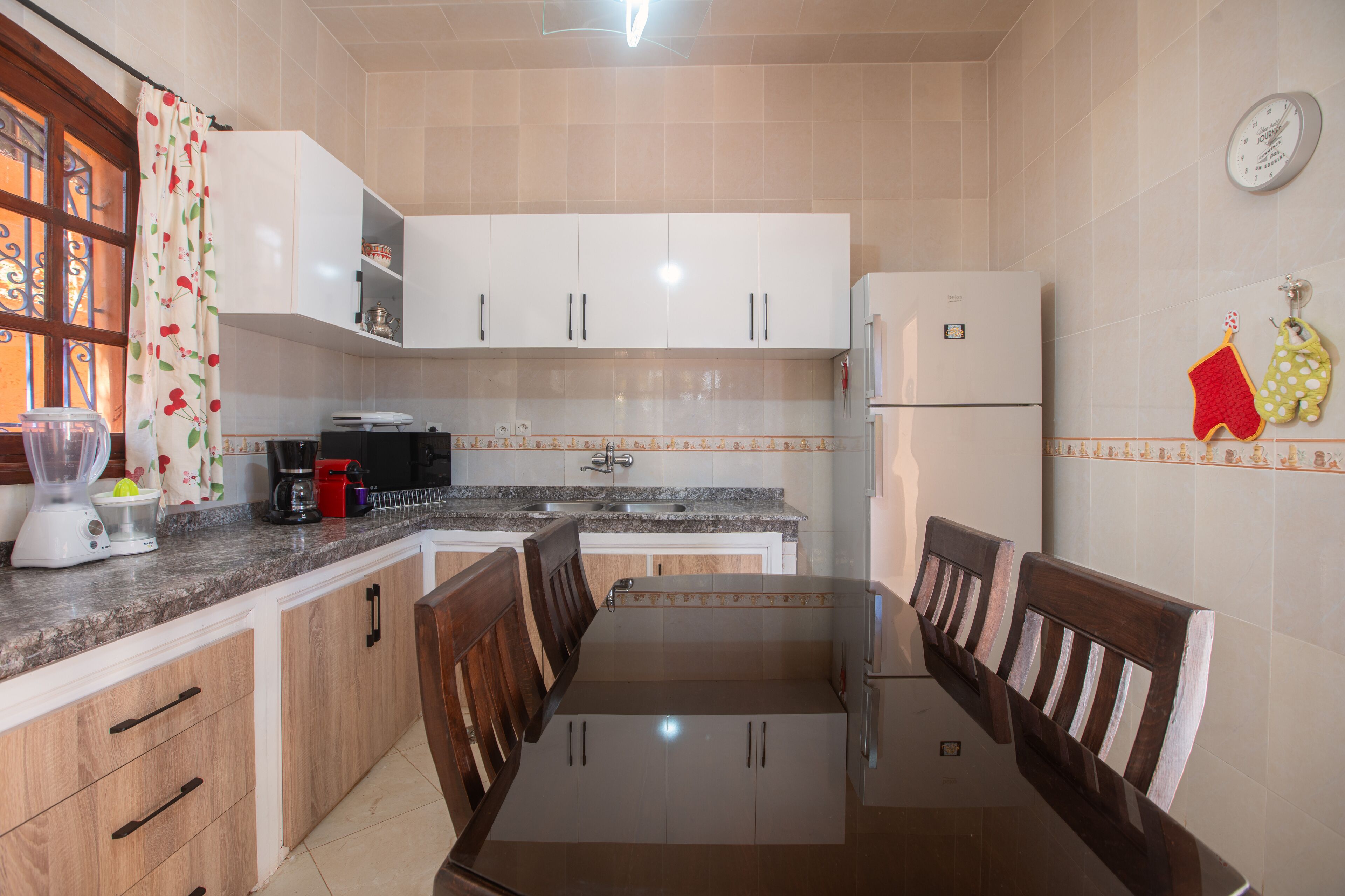 A kitchen with white cabinets, a granite countertop, a dining table with chairs, a refrigerator, and a clock on the wall.