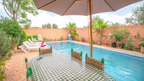Une piscine avec des chaises longues, une table à carreaux et un grand parasol.
