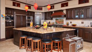 A modern kitchen with dark wood cabinets, a large island with stools, stainless steel appliances, and decorative hanging lights. - 7 Bed Waterfront W/Rooftop pool & hot tub stunning 360-degree bay & beach view (Newport Beach)