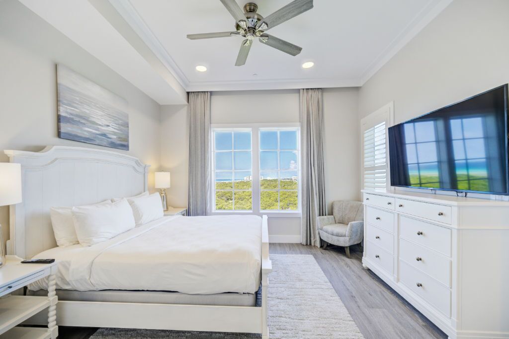 A hotel bedroom with a large bed, a ceiling fan, a TV, a window with curtains, and a view of the outdoors.