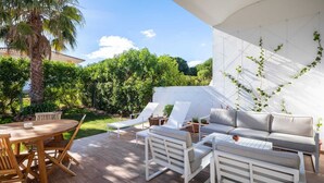 Um pátio com uma mesa redonda de madeira, cadeiras brancas, um sofá cinzento e um guarda-sol branco.