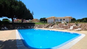 Piscina com espreguiçadeiras, um edifício ao fundo e um caminho de pedra.