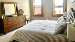 Une chambre à coucher comprenant un lit, une commode, un miroir et deux fenêtres équipées de stores.