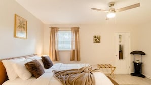 Une chambre à coucher comprenant un lit, un ventilateur de plafond, une fenêtre avec des rideaux, une porte et une table de chevet.