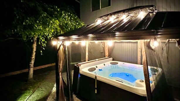 Een hottub onder een overdekte ruimte met lichtjes, omringd door een grindpad en een boom.
