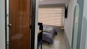 A narrow hallway with a wooden door on the left, a bed with a headboard and pillows in the center, and a small table with a chair. - Apartment 10 Min Airport F

 (Bogotá)