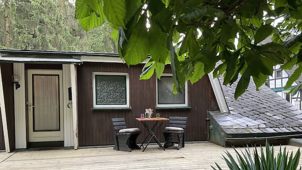 A wooden deck with a table and two chairs, a door, and two windows. - Wohnung in Idyllischem Örtchen (Meschede)