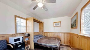 Quarto compacto com cama, pequena cozinha, ventilador de teto e janela com cortinas.