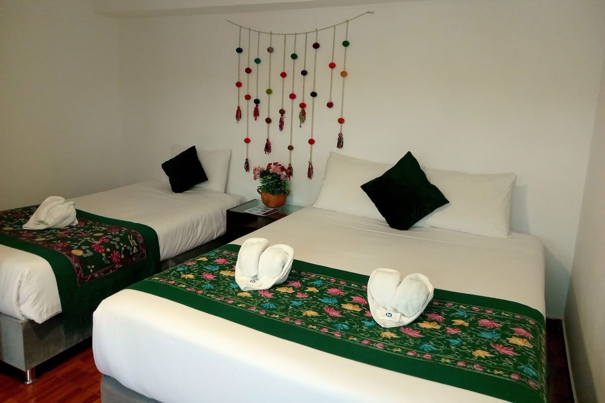 Quarto de hotel com duas camas de solteiro, cada uma com uma almofada branca e um edredom verde com padrão floral. Há uma pequena mesa com uma planta e um ornamento pendular decorativo entre as camas.