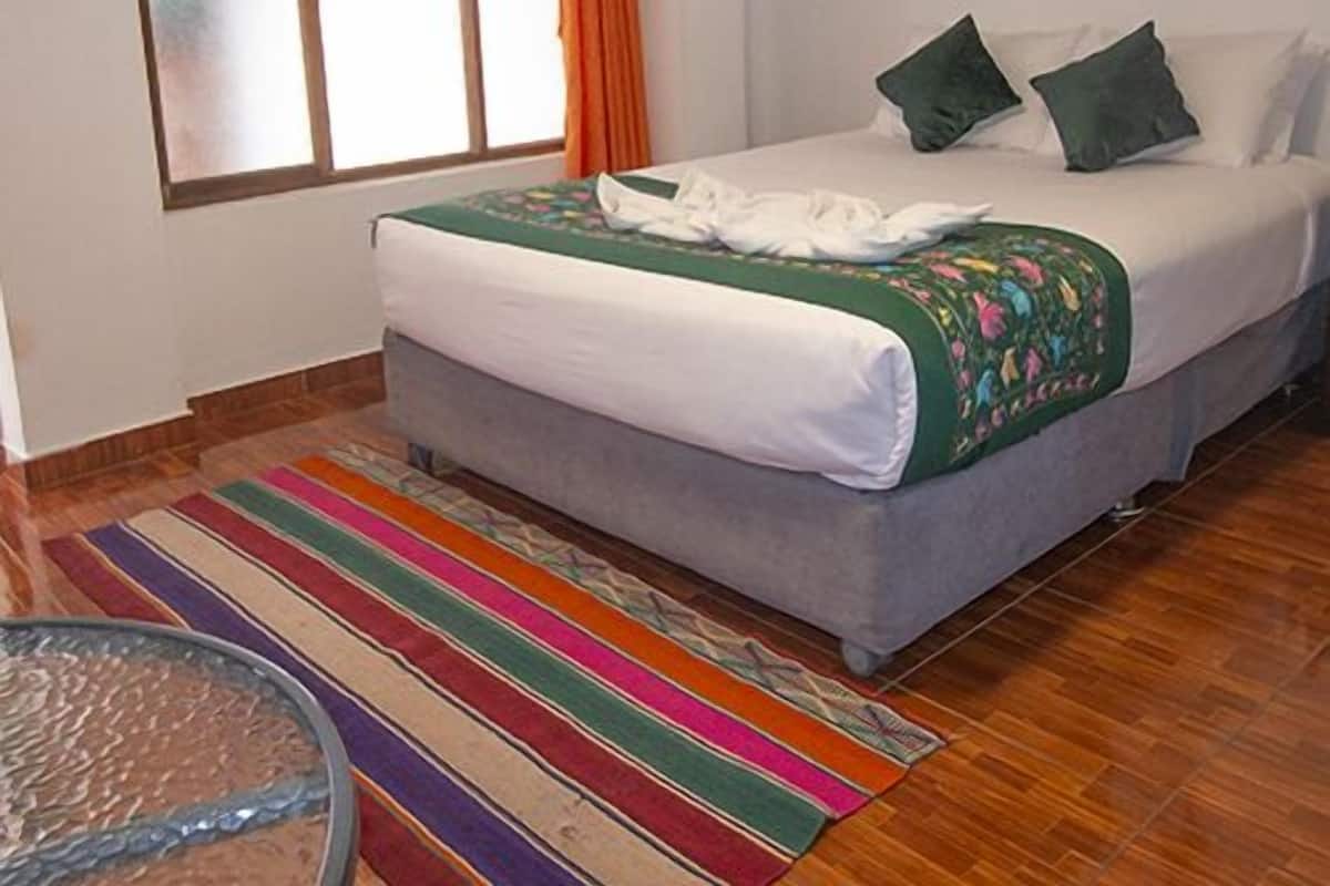 Um quarto com cama, janela com cortinas, um adorno pendurado e um tapete colorido.