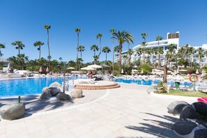 A resort pool area with palm trees, lounge chairs, and a large building in the background.