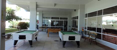 A covered outdoor area with two pool tables, a foosball table, and a seating bench.