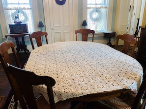 Una mesa redonda con mantel de encaje, sillas de madera, una mesita con un radio y cortinas de encaje.