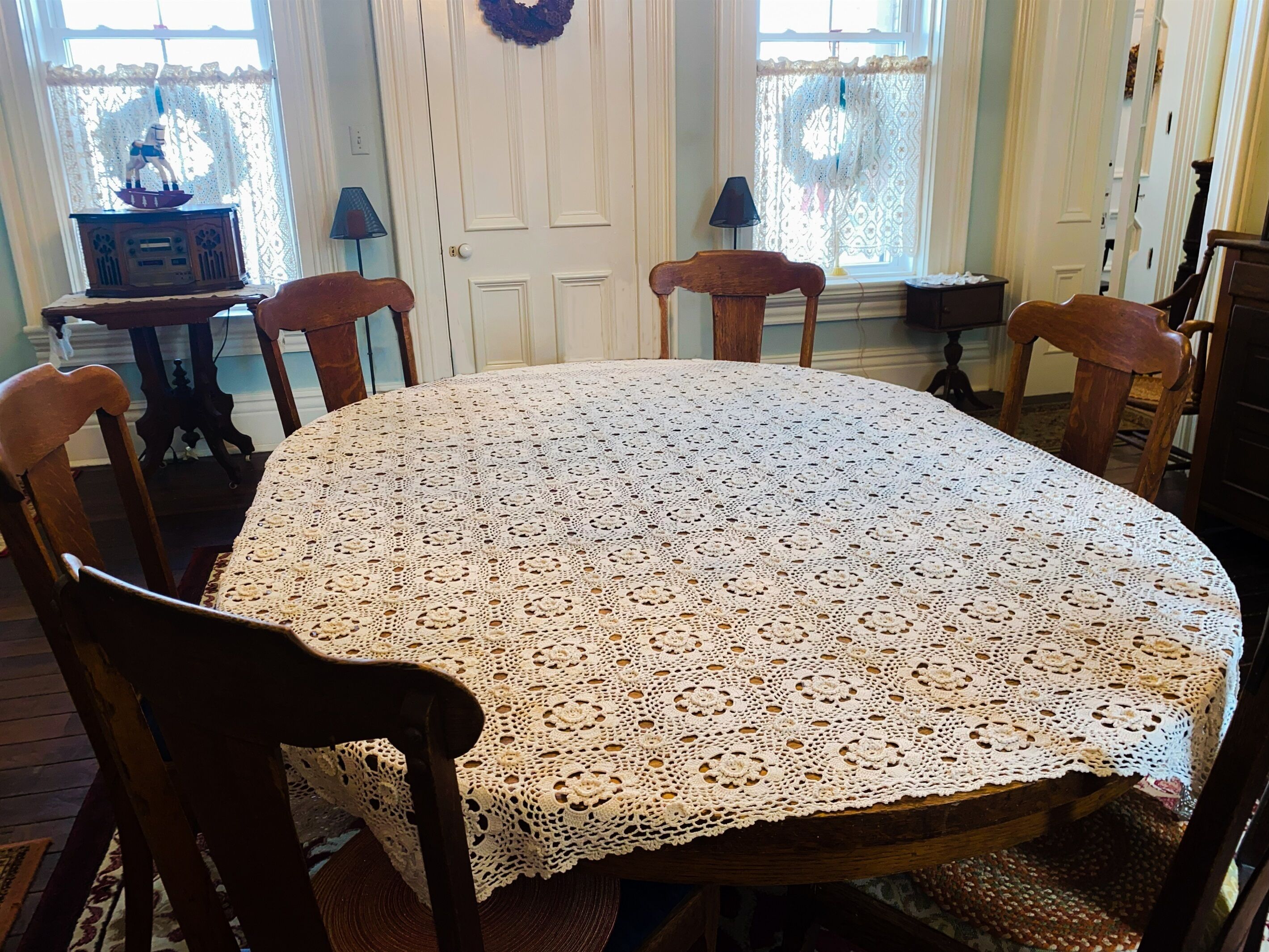 A round table with a lace tablecloth, wooden chairs, a small table with a radio, and lace curtains.