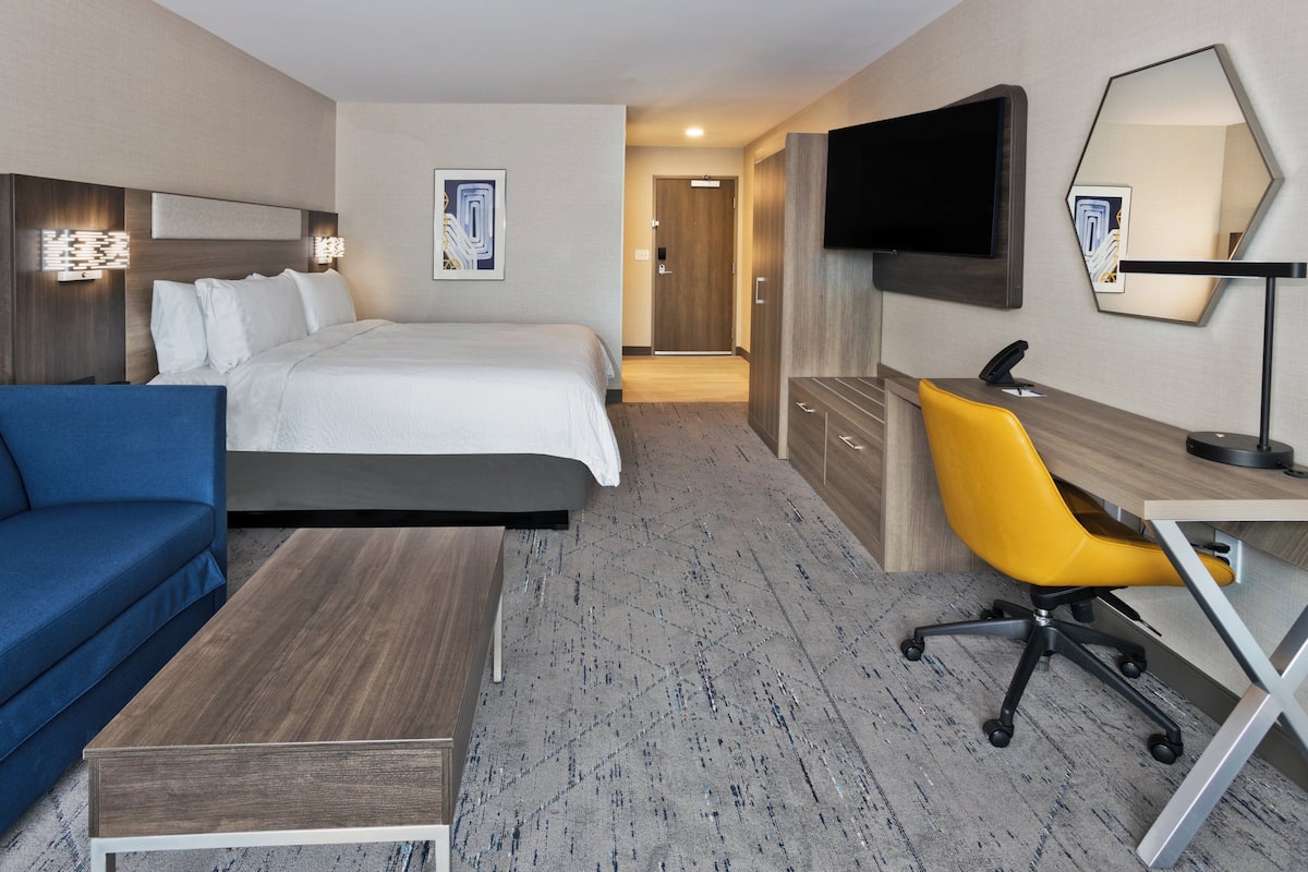 A hotel room with a bed, a blue sofa, a desk with a chair, a flat-screen TV, and a mirror.