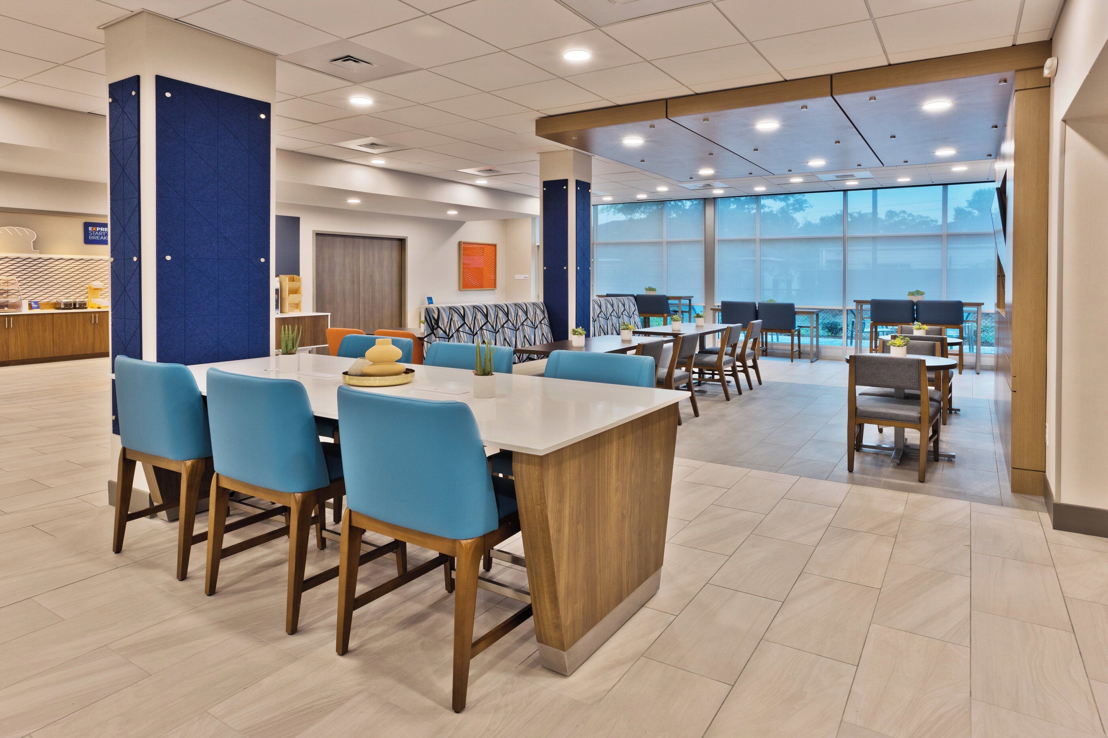 A modern hotel lobby with a central dining area featuring blue chairs and a wooden table, surrounded by large windows offering a view of the outdoors.