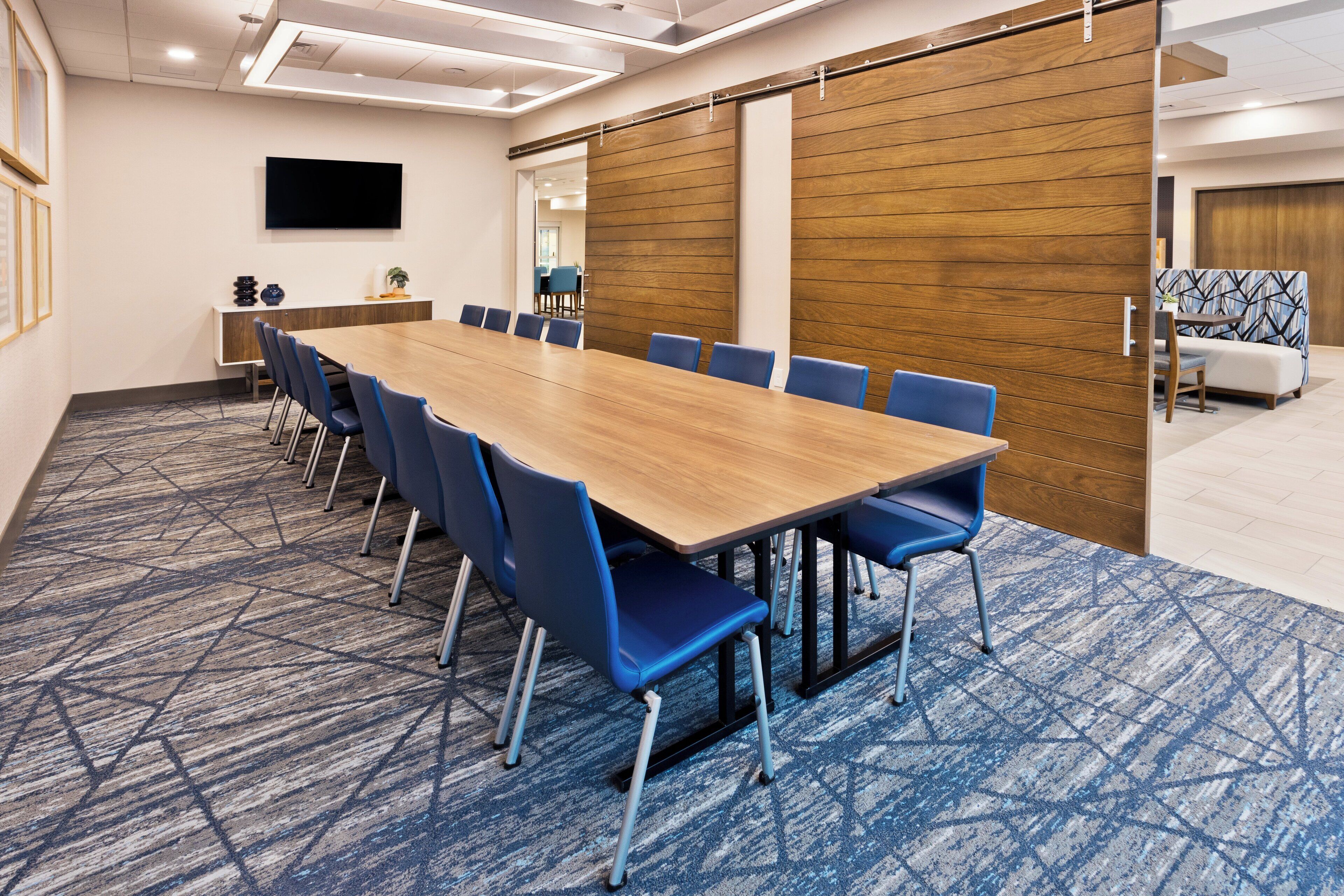 A conference room with a long table, blue chairs, a flat-screen TV, and a wooden partition wall.