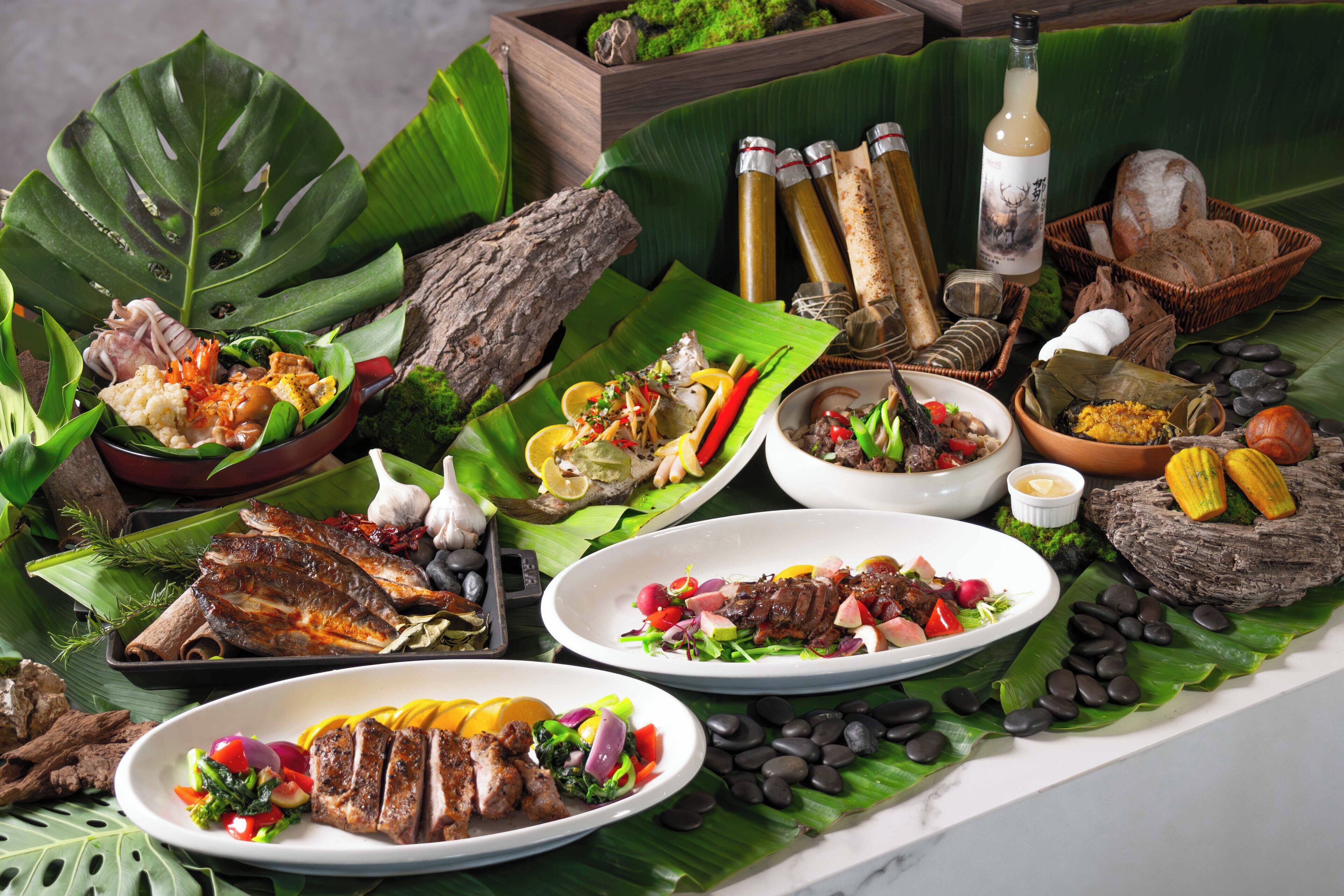 Una variedad de platos y alimentos presentados sobre una mesa con temática tropical, incluyendo frutas, verduras y carnes, con un fondo de hojas verdes y elementos naturales.