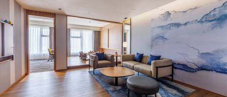 A modern living room with a sofa, coffee table, and ottoman, a large abstract painting on the wall, and a view into the dining area.